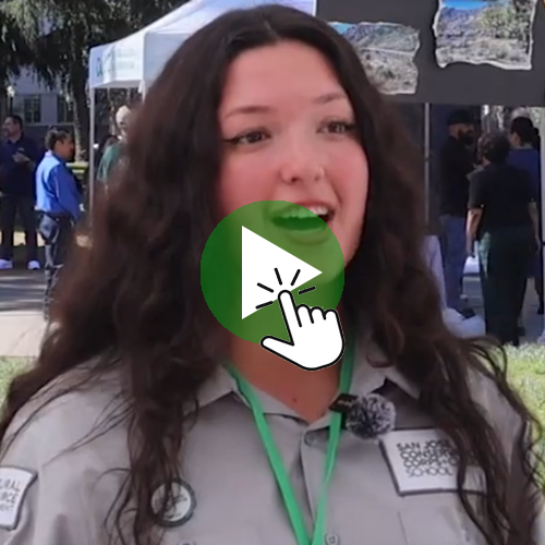A person wearing an outdoor‑event uniform stands in front of booths and display boards, with a green play‑button icon and hand‑cursor graphic overlaid in the center. Persona con uniforme en un evento al aire libre, con un ícono de botón de reproducción y un cursor superpuesto al centro.