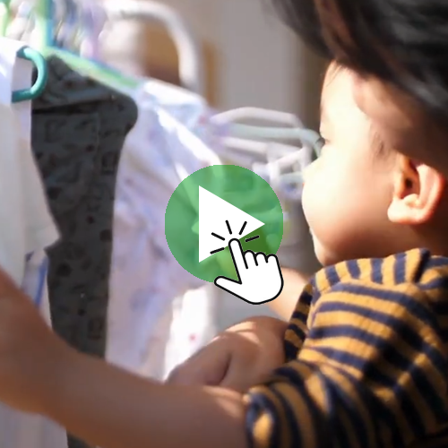 A child reaches toward clothing hanging on a rack, with a green play‑button icon and hand‑cursor graphic overlaid in the center. Niña o niño alcanzando ropa en un estante, con un ícono de botón de reproducción y un cursor superpuesto al centro.